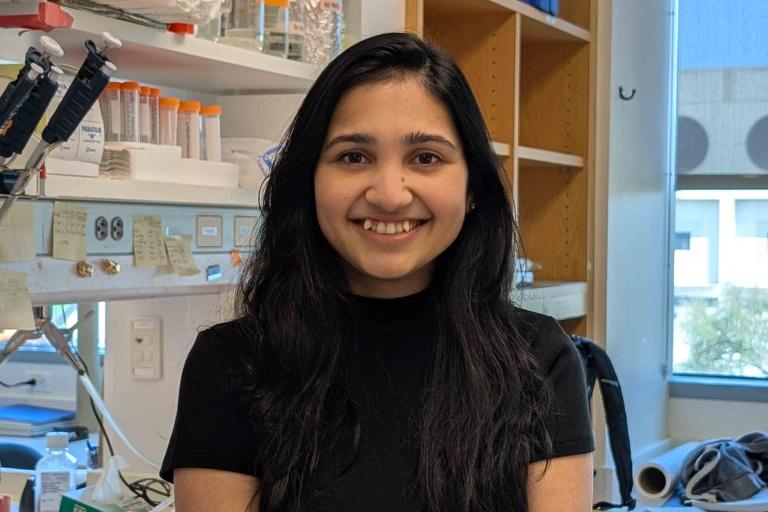 Smiling woman with long black hair and black shirt stands in front of a lab bench. 
