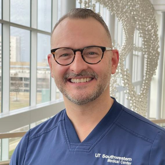 Smiling man with fair hair, wearing dark rimmed glasses and blue UT Southwestern Medical Center scrubs.