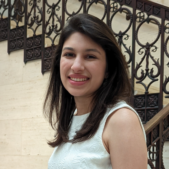 Smiling woman with long brown hair, standing beside a staircase.
