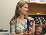 Smiling woman with long light brown hair, wearing a striped shirt and black pants, standing next to a bookcase, holding a bag of peanuts. Two laughing women are seated next to her.
