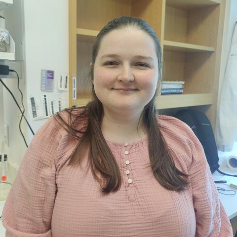 Woman with brown hair and a pink shirt stands in front of a lab desk