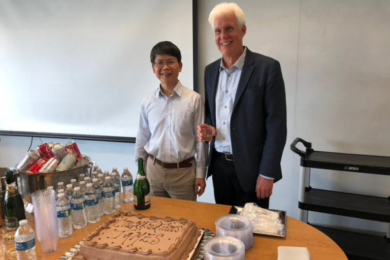 Two smiling men standing behind a table laden with drinks, paper goods, and a cake that says, Congratulations, James!