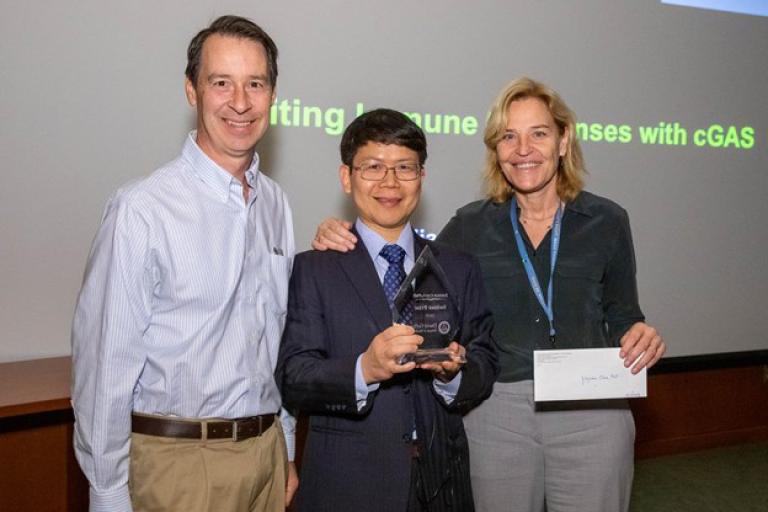 Smiling man with dark hair and glasses,  holding a tear-drop shaped award, flanked by a tall smiling man with short dark hair and a smiling woman with shoulder-length blond hair.