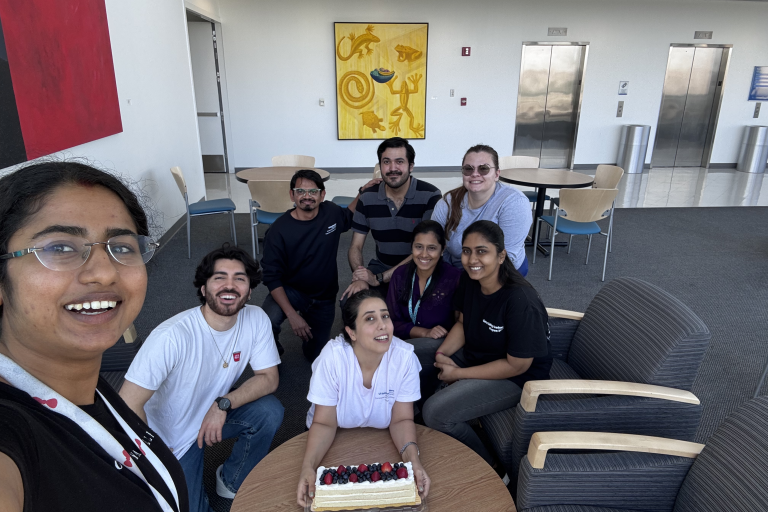 A group of eight people pose for a photo in a hotel setting with one holding a cake.