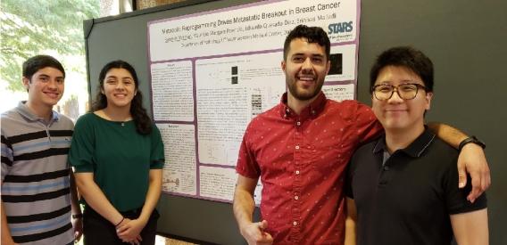 Four lab members pose for the camera in front of poster presentation