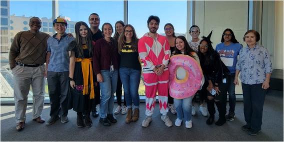 14 lab team members in front of a large window, some in costumes.