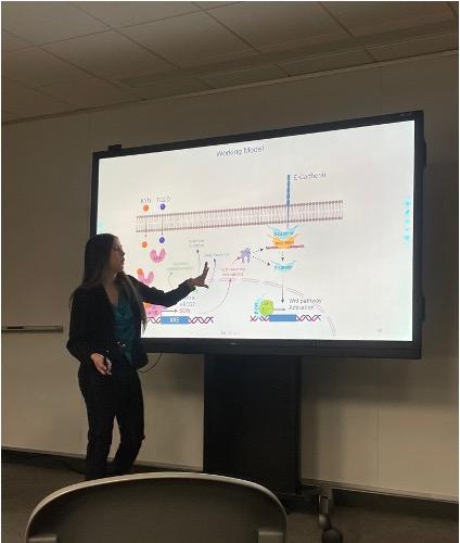 A woman stands in front of a screen speaking to an unseen audience.
