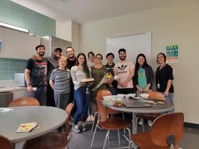 The lab team assembled in a break room, two women in front holding a cake and flowers.