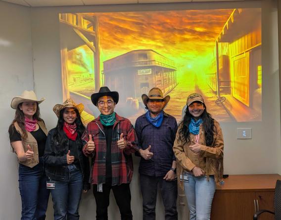 Five individuals in cowboy hats and western attire in front of a western movie style image. 