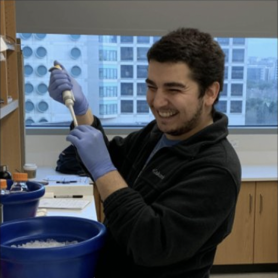 Smiling man with dark hair and trim dark facial hair, wearing dark clothing and blue latex gloves, holding a dropper over a blue bucket.