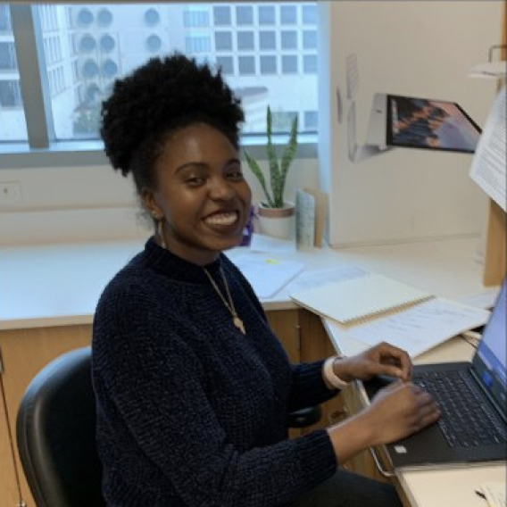 Smiling woman with a black pony tail/bun wearing a dark sweater, seated at a desk working on a laptop computer.