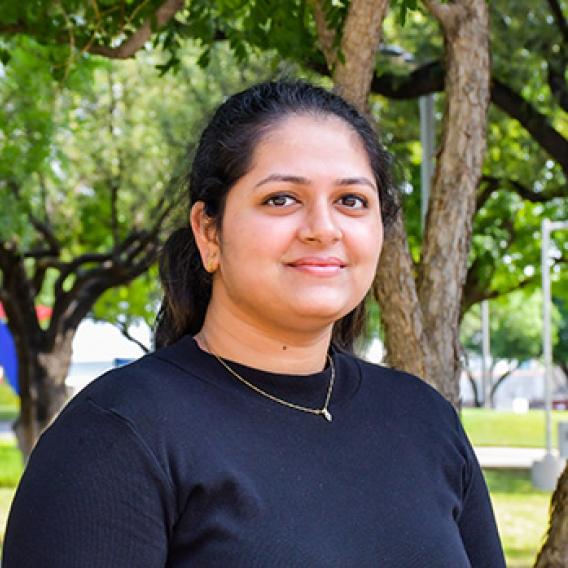 Woman with dark hair pulled back in a pony tail wearing a blue shirt standing outside among trees.