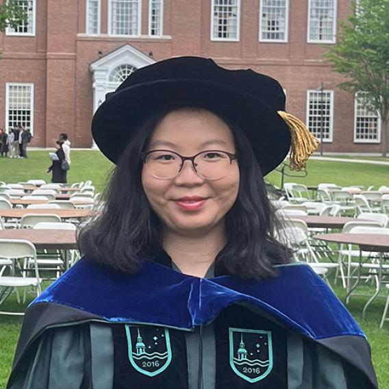 Smiling woman with long dark hair, wearing scholarly robes and glasses.