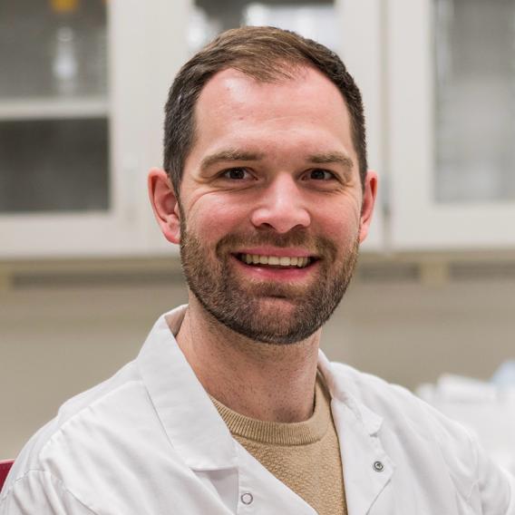 Smiling man with dark hair, beard, and mustache, wearing a white lab coat, posing in a lab.