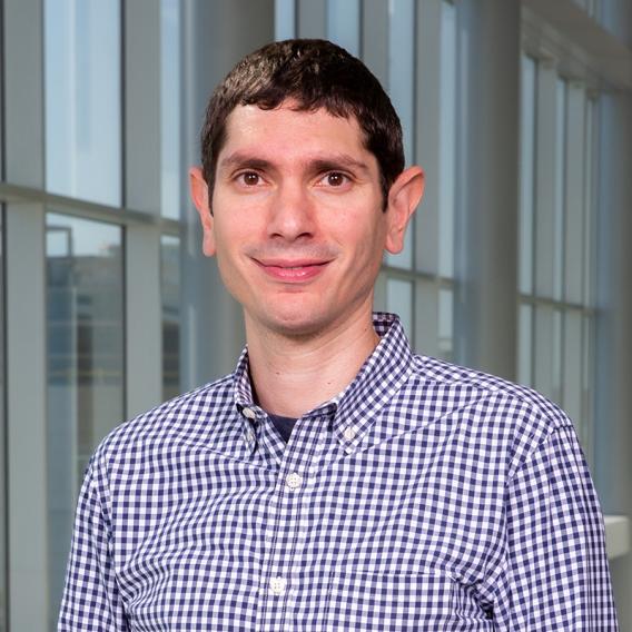 Smiling man with short, dark hair, standing outside wearing a blue-checked shirt.