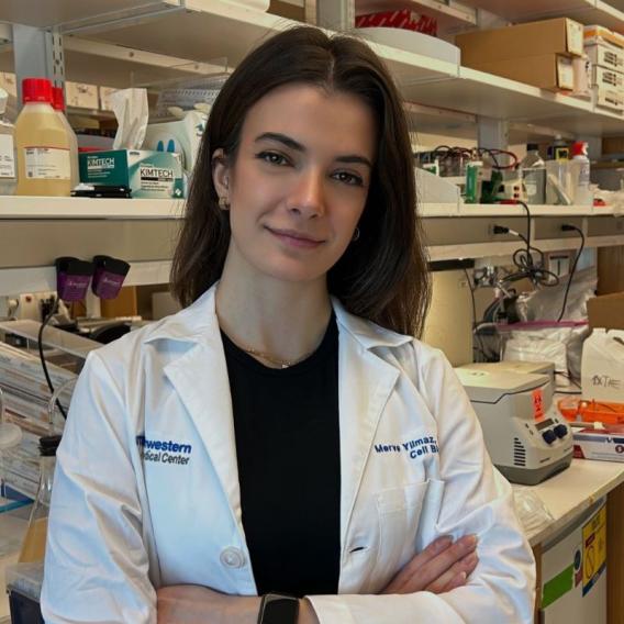 young woman with long dark hair wearing white lab coat stands with arms crossed in front of shelves with lab supplies and equipement