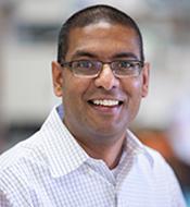 Smiling man with closely cropped dark hair, wearing a white checked shirt and half-rimmed glasses.