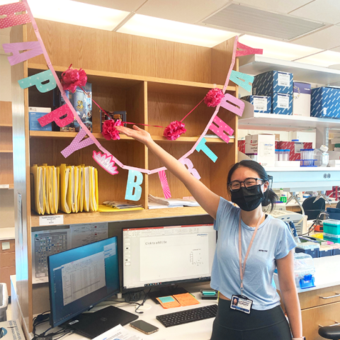 Maggie wearing face mask, standing with "Happy Birthday" banner