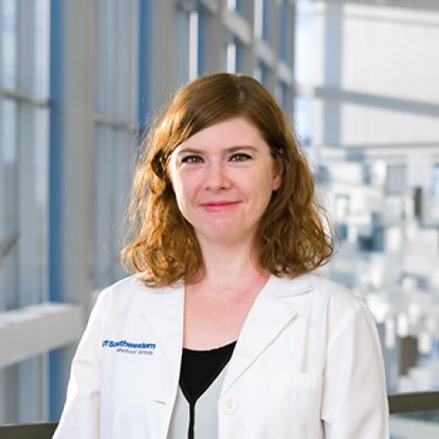 Woman with auburn shoulder-length hair wearing a lab coat, standing in front of large industrial windows.