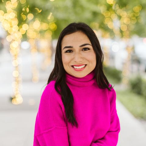 Smiling woman with long dark hair, wearing a pink turtle-neck sweater, outdoors.