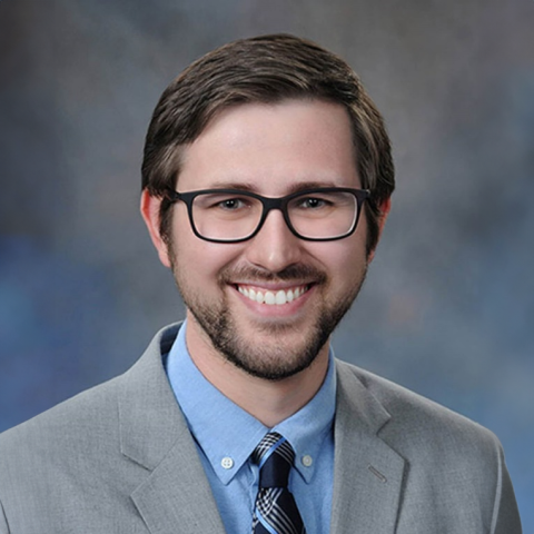 Dr. Alexander Tatara wearing a grey suit, blue shirt and striped tie on a mottled blue-grey background