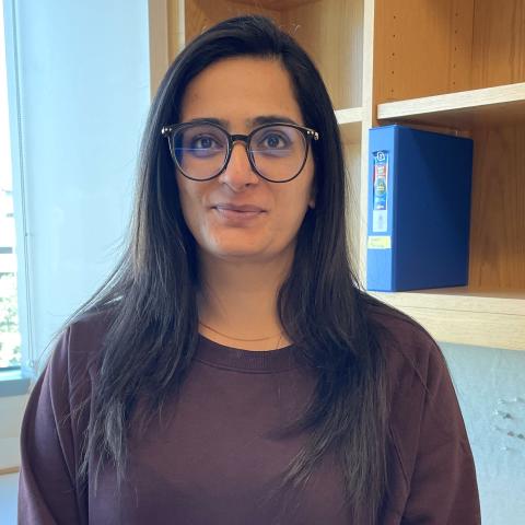 A woman with long dark hair, a dark sweater, and glasses in front of a lab bookshelf. 